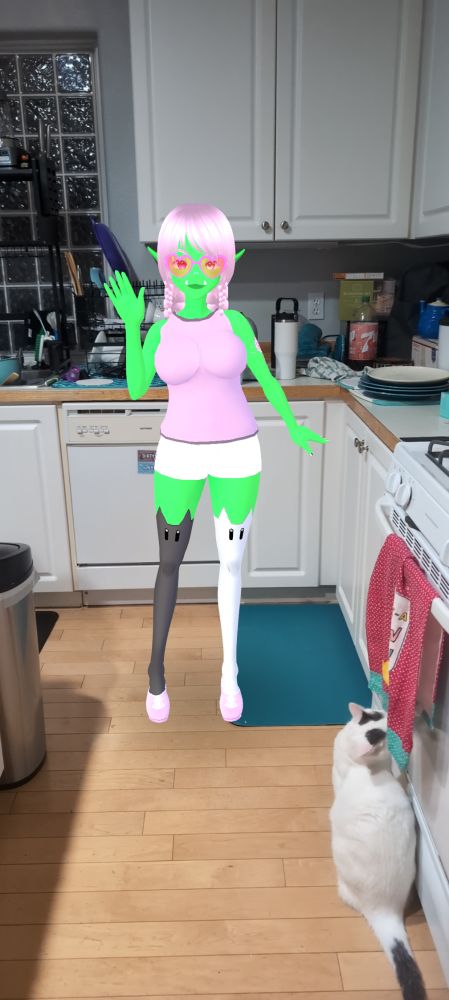 A green skinned orc with pink hair tied up in twin braids waves and smiles at you from a corner of a kitchen, where there are stacked dirty dishes and a nosey white cat who is sniffing a towel on the oven door.