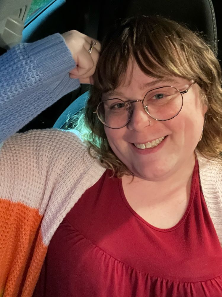 lily, wearing a red shirt and multi colored cardigan, sitting in the driver’s seat of her car