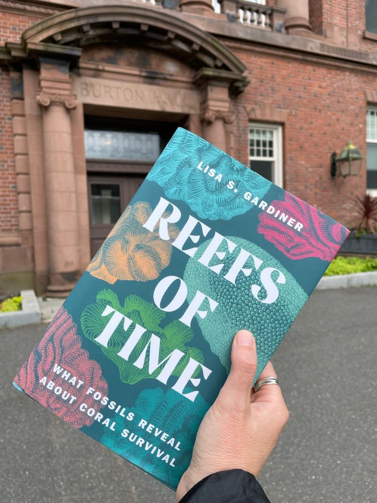 Hand holding up a book titled _Reefs of Time: What fossils reveal about coral survival_ in front of a brick academic building with “Burton Hall” chiseled above the door.