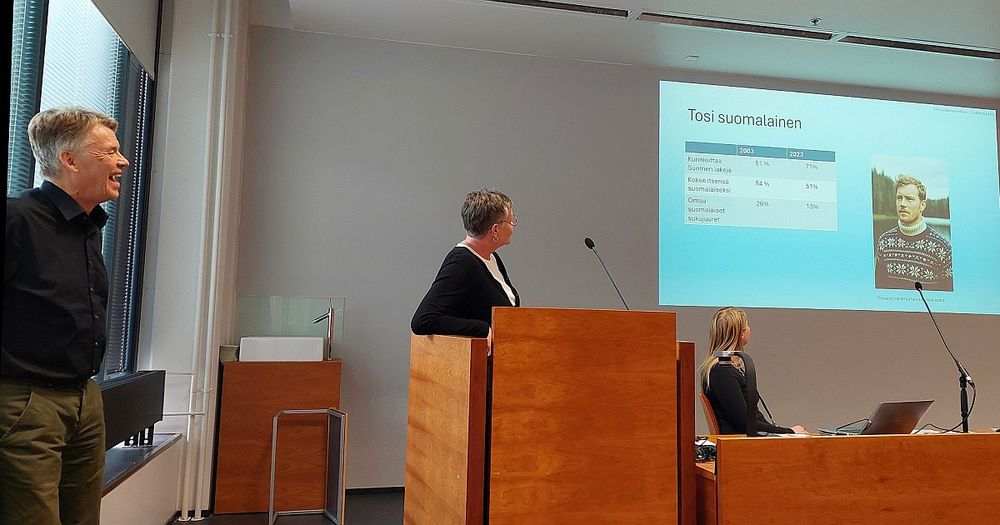 Kolme ihmistä luentosalissa, katsovat ruudulle heijastettua kuvaa.