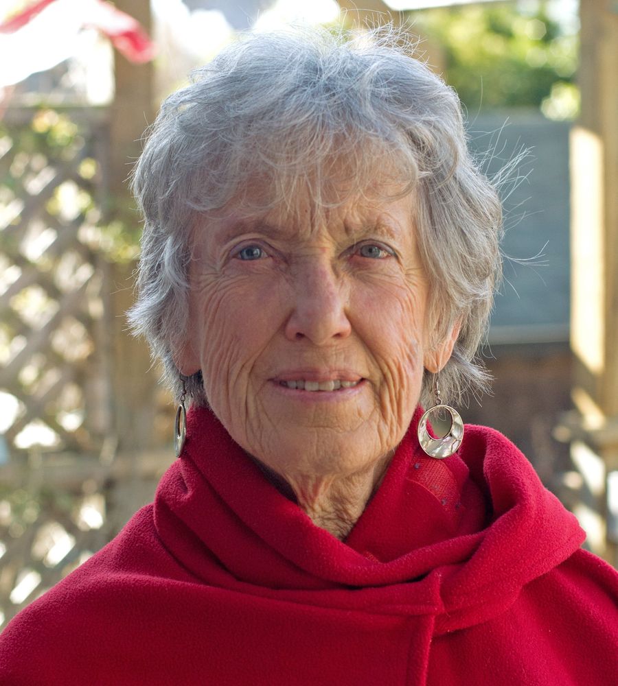 A headshot of Joanna Macy in her later years. She has grey hair and mother-of-pearl earrings, with a red cloak. Picture is from https://believe.earth/en/