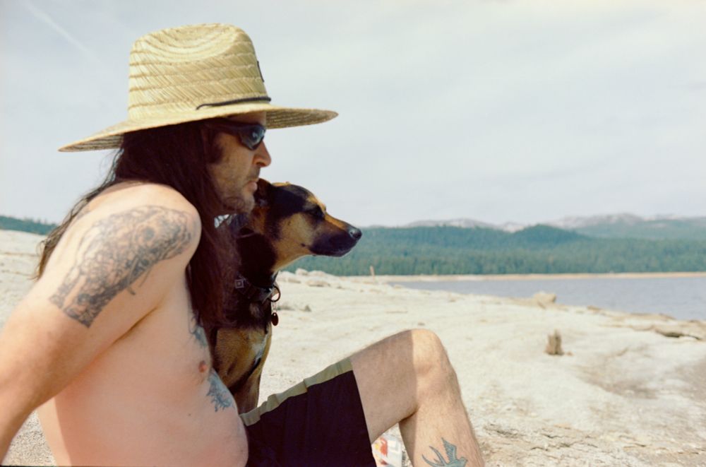 A tattooed man with long dark hair wearing a sun hat sits in swim shorts next to a black and tan large dog, both looking out onto a lake in the sun. 