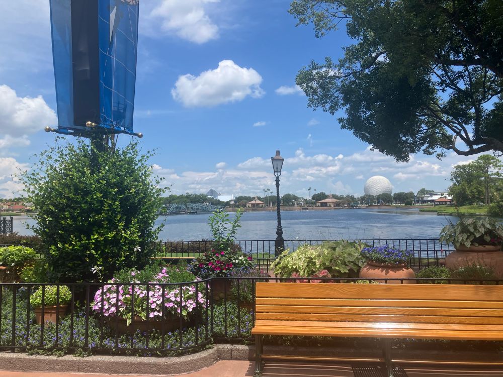 Purple flowers and a wooden bench in the foreground; a lake and the EPCOT geodesic sphere in the background. A blue sky above.