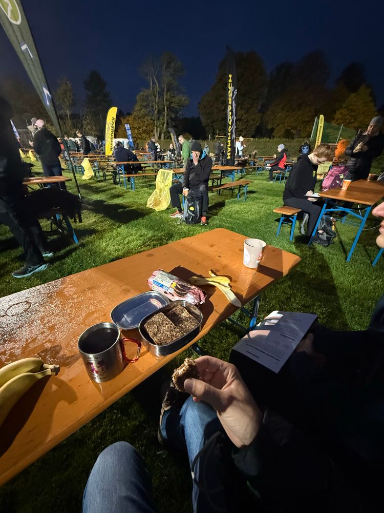 Foto einer frühen Morgenstimmung bei einer Outdoor-Veranstaltung. Menschen sitzen in Jacken und Mützen an Bierbänken auf einer nassen Wiese. Im Vordergrund liegt auf einem Holztisch ein Frühstück mit Banane, Müsliriegel, Brotdose mit Brot und einem Metallbecher. Eine Person hält ein Stück Brot in der Hand. Im Hintergrund sind Fahnen und weitere Teilnehmende zu sehen.