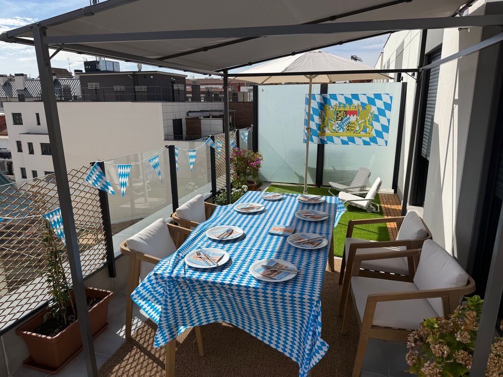 Ein gedeckter Terrassentisch. Die Tischdecke mit bayrischen Rauten, am Balkongeländer hängen weiß blaue Wimpel, im Hintergrund die bayrische Landesflagge an der Wand. 