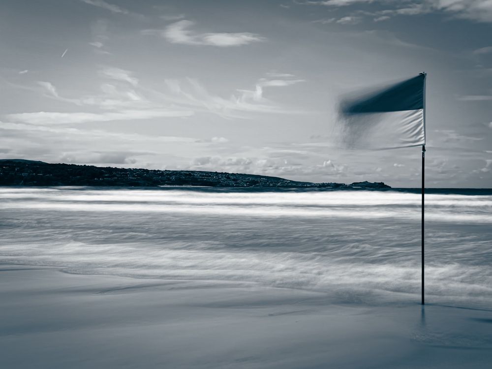 A photo taken from Hayley beach in Cornwall looking southwest towards St Ives in the distance. The photo is converted to black and white using Adobe Lightroom and then cooled so has a blue cast to it. In the photo on the right hand side is the lifeguard’s red/yellow flag. The photo has been taken with a iPhone and set to long exposure therefore making the sea and waves milky, and the red/yellow flag blowing in the wind is also slightly blurred.