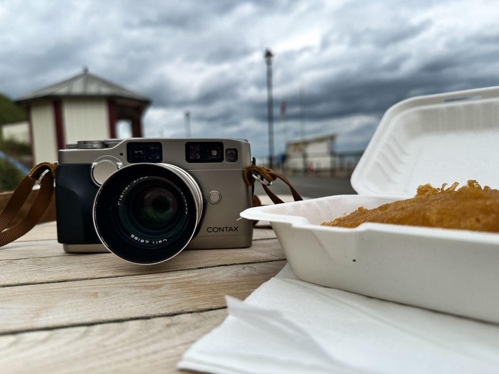 Contax G2 camera on a table next to a paper tray for fish and chips, containing only a battered cod. Fish and Chips by the seaside, Saltburn-by-the-Sea, Cleveland. 