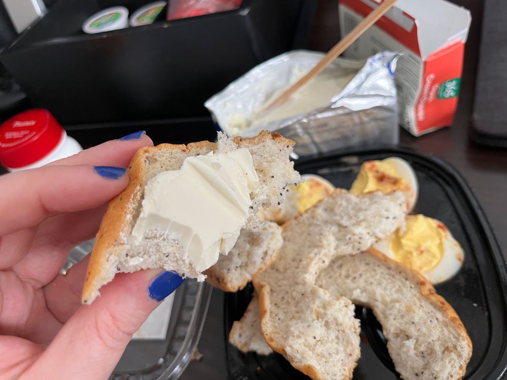 A hand with blue nail polish holds up a quarter of a bagel with poorly spread cream cheese. In the background is packaged devilled egs and a coffee stirrer stuck into a cream cheese block.