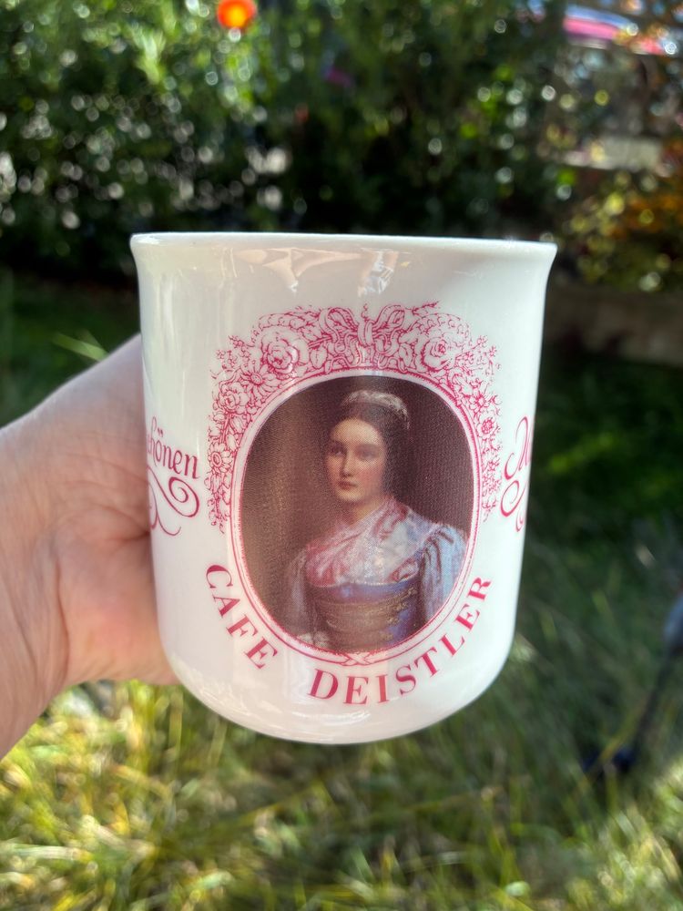 Vor verschwommenem Gartenhintergrund eine Hand mit einer weißen Tasse mit rosa Aufdruck von Café Deistler, mittig das Porträt von Helene Sedlmayr aus der Schönheitengalerie von König Ludwig I