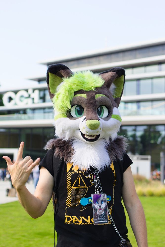 Photo of the fursuiter Fara. The fursuiter only wears a furry head of a fox with green, brown and white colors while showing the sign of the horns. In the background the Convention Center Hamburg (CCH) is visible.
