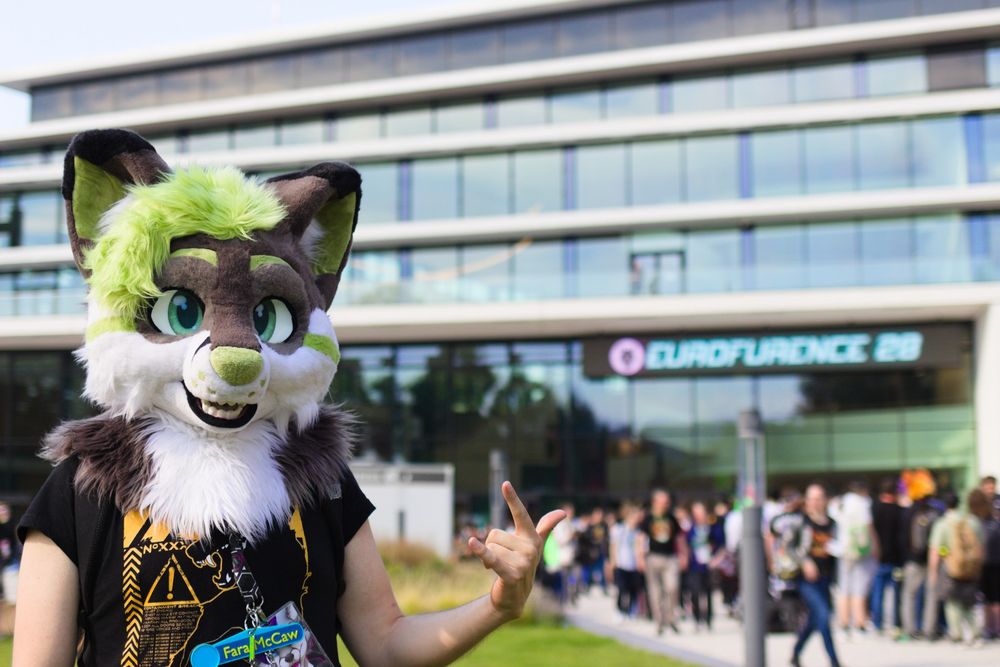 Photo of the same fursuiter, but from a different angle. In the background there is a queue of furries without fursuits waiting to register for the Eurofurence. The sign of horns is now shown with the other hand.