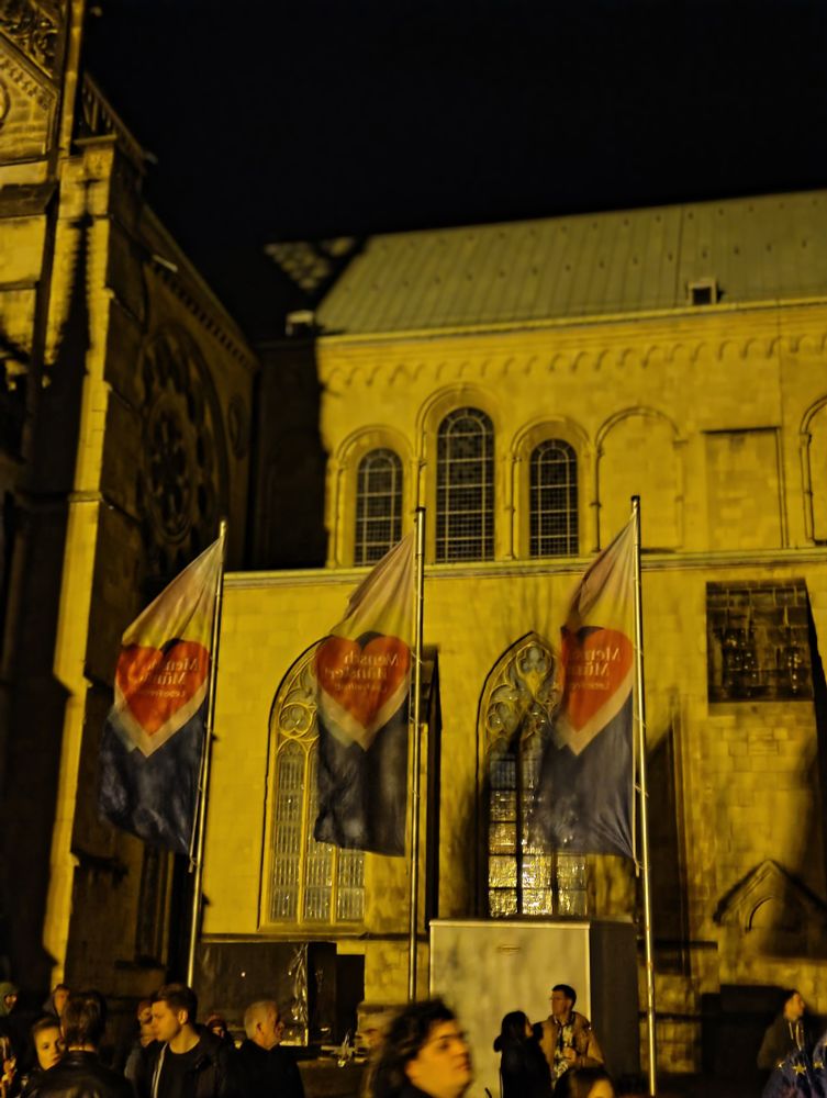 Schriftzug aufFahnen vor einem Dom, auf dessen platz eine Demo stattfindet. Die Fahnen sind an die LGBTQ+ Farben angelehnt. Schriftzug nicht klar lesbar, sry.