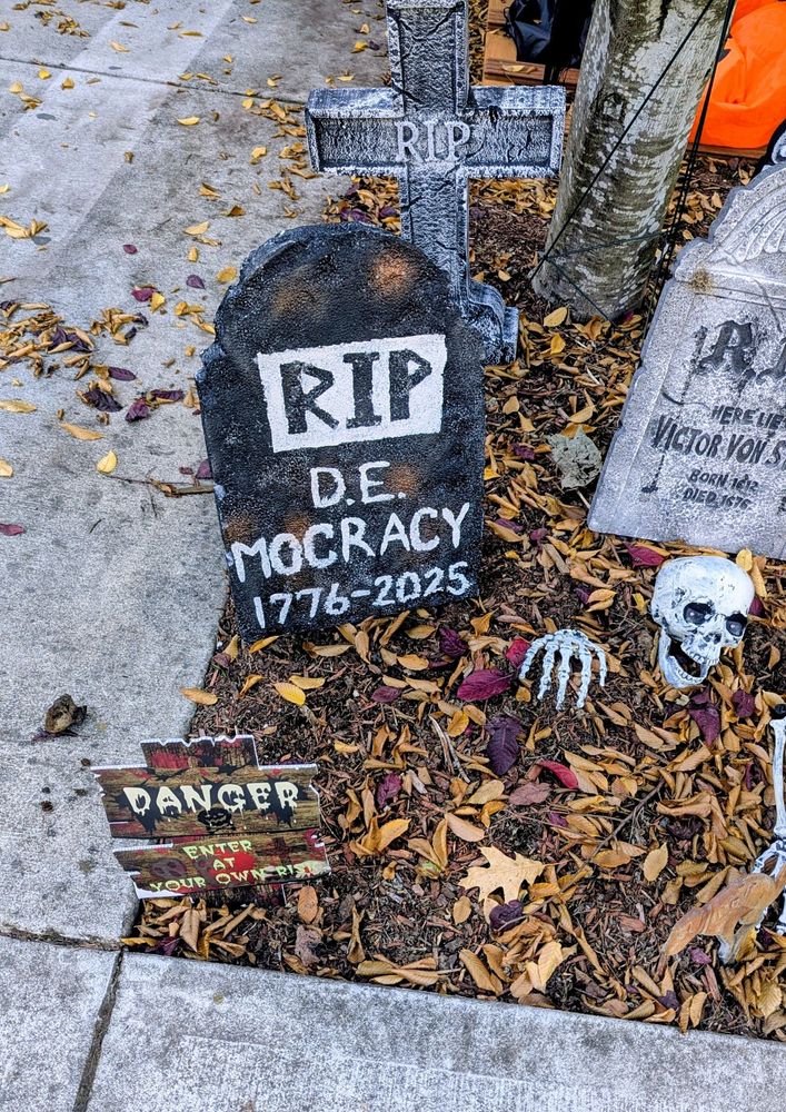 A Halloween yard decoration, a tombstone reading RIP D.E. Mocracy, 1776-2025. A skeleton is coming up out of the grave and there are other Halloween tombstones nearby.