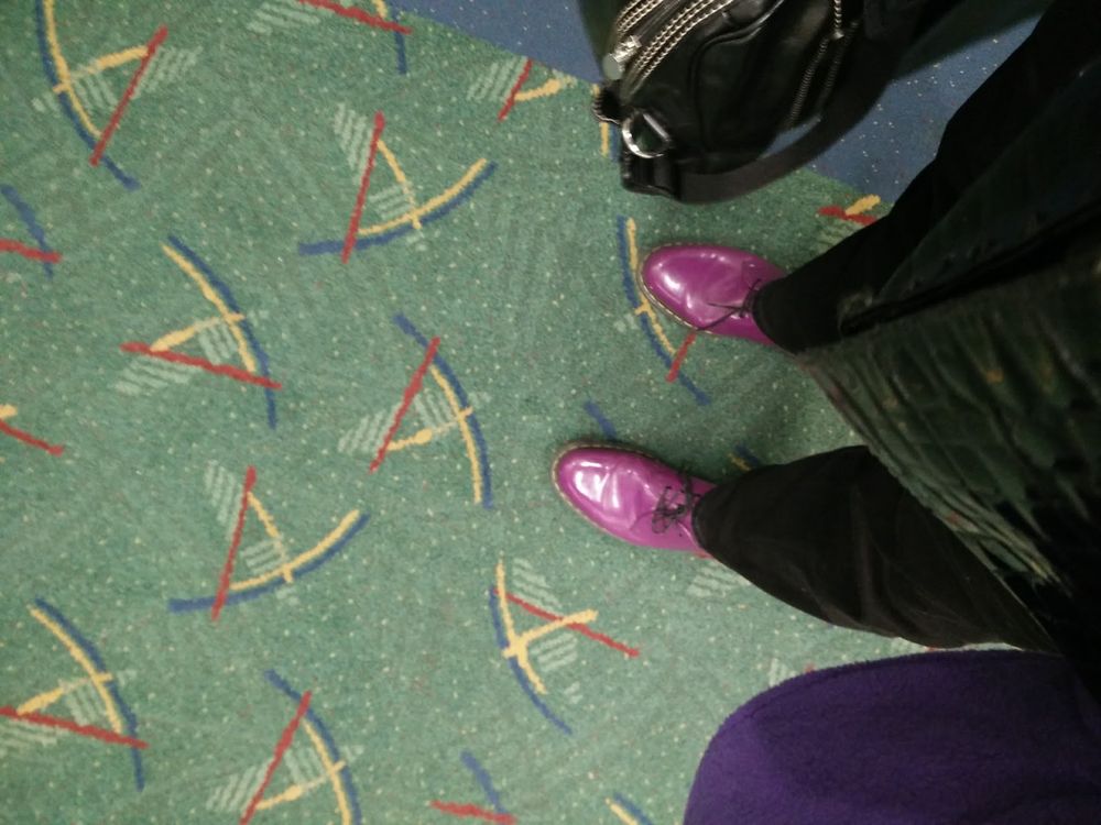 Photo of a person's feet in purple patent leather Doc Martens standing on the famous green patterned carpet at PDX, Portland International Airport.