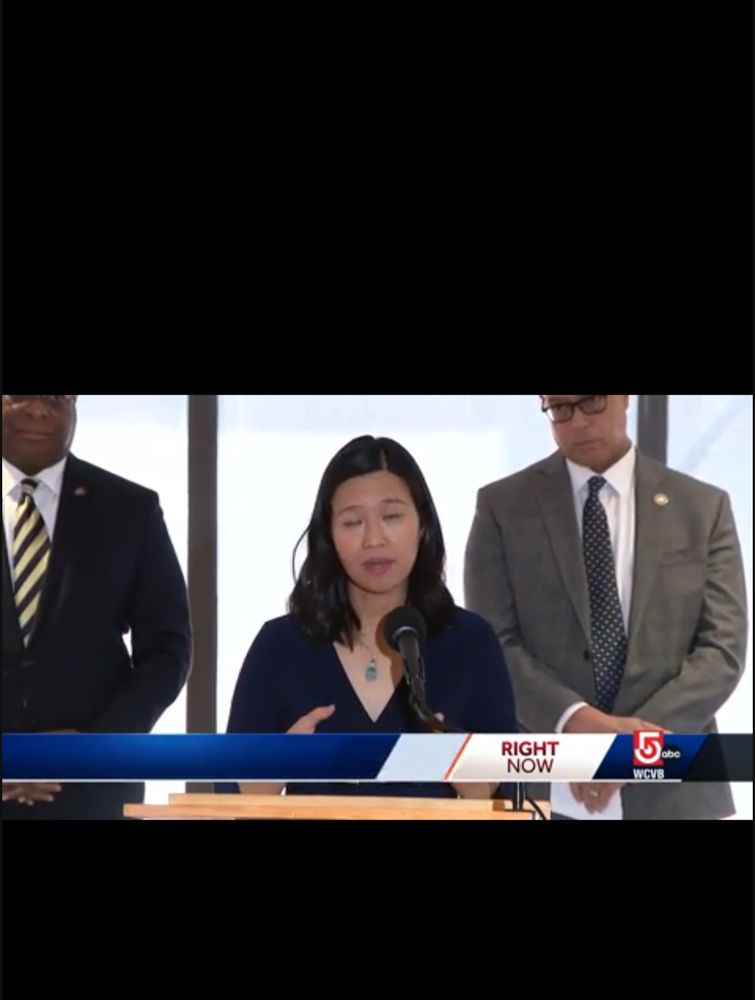 Screencap of a video of Mayor Michelle Wu in front of all white windows, with two men in suits partially cut off in the background