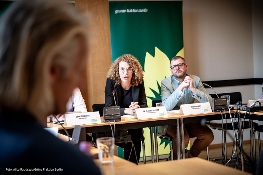 Foto von Claudia Leistner und Werner Graf, die zusammen an einem Tisch sitzen, im Hintergrund ein grünes Banner. Im Vordergrund eine Person im Anschnitt, die den beiden zuhört.