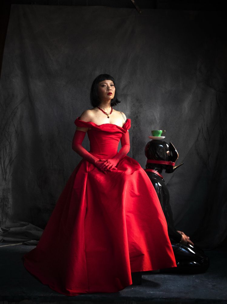 An Asian femme sits on a platform in a red ballgown with hands folded in front of her, chin tilted up. To her side is a black latex clad figure seated on the floor, facing away, with a red leather strap fastening a glossy black stiletto heeled shoe to their face, and a bright green teacup and saucer placed atop their head. The background is a dark grey backdrop. 