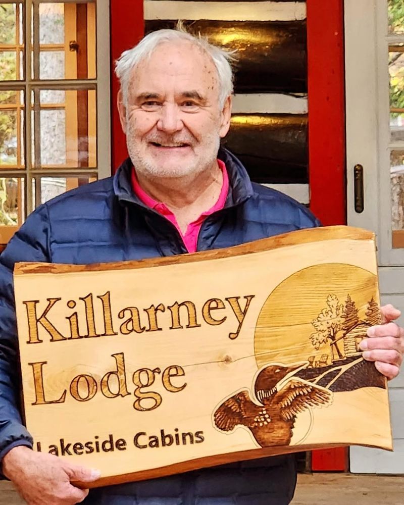 Miglin holding up a wooden plaque that says "Killarney Lodge Lakeside Cabins"