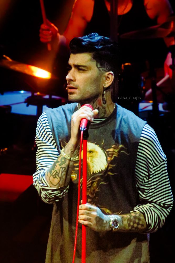 Zayn on stage wearing a harley davidson shirt with an eagle on his chest and striped sleeves. He's holding on to the mic and looking out into the crowd