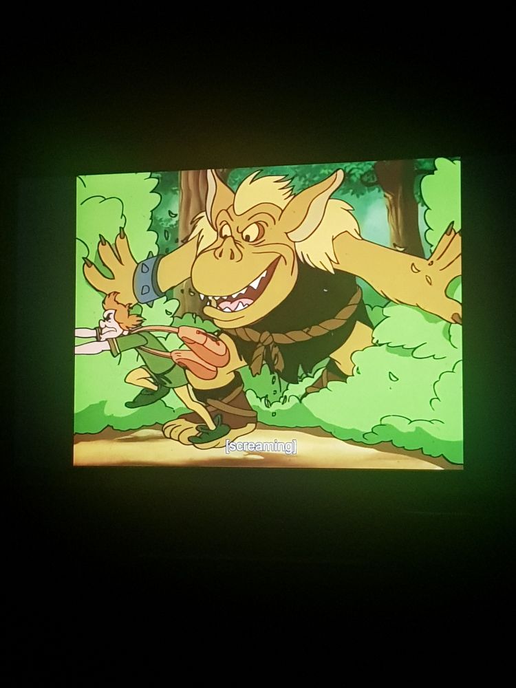 An ogre showersy from behind bushes as a man in medieval everyday clothes runs away. They're in a forest.