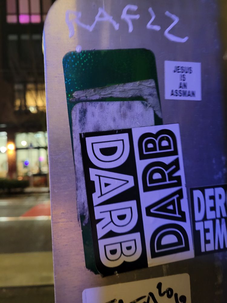 Stickers on the back of a sign.
One reads "Jesus is an assman"