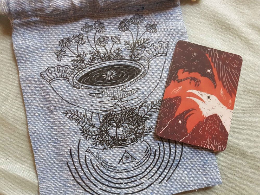 Light blue dice back with a black, simple lines print of hands holding a cup of tea surrounded by herbs, and an art card of black and white ravens on a red background