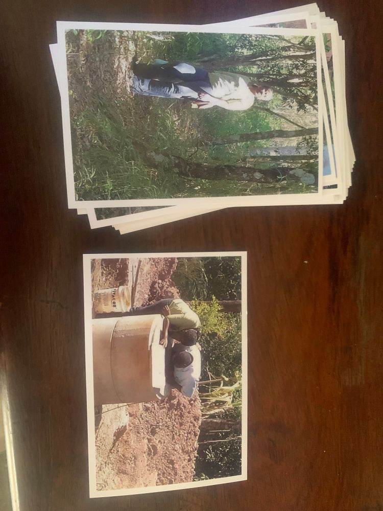 Foto colorida, uma foto de duas fotos sobre uma mesa de madeira, em uma das fotos uma mulher de aparentemente 40 anos em uma mata fechada, ela é negra está de cabelos presos e usa uma blusa e calça. Na outra foto um casal de pessoas negras, ele e ela, olha para dentro de um poço d’agua inacabado, em segundo plano um terreno
