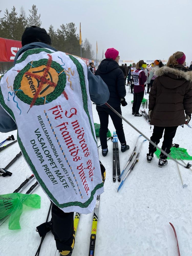 Bilden visar skidåkare som väntar på att få starta i Vasaloppet. Över ryggen på den som är närmast bild hänger ett tygstycke. På tyget står det ”I mödrars spår för framtidsvintrar. Vasaloppet måste dumpa Preem!” företaget Preem logga finns också på tyget. Den är överkorsad med rött.
