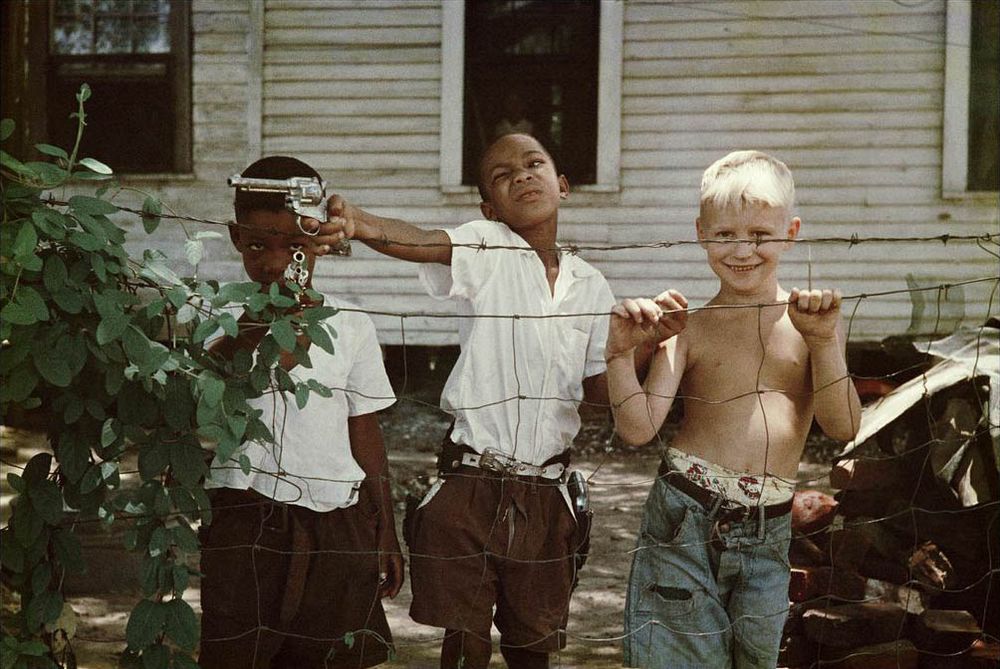 A color photograph of three young boys standing in front of a chicken wire fence in the yard of a white wooden house, somewhat grubby. Two are Black, one is white and quite blond; the Black boys are wearing matching  white shirts and dark shorts. The white boy is shirtless and wearing jeans, his underwear peeking out. The two Black boys are playing with cap pistols, one pointing his off-camera to the side, the other pointing it at the photographer. The white boy is holding onto the fense and looking at the photographer, grinning.