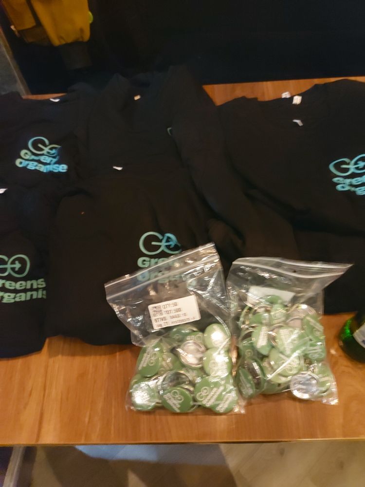 Black t shirts with a green logo reading 'Greens Organise' are piled on a wooden surface. In front of them are two polythene bags full of green and silver badges.