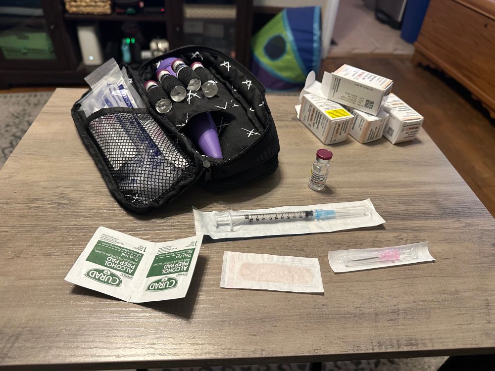 The photo is of HRT supplies on top of a TV table tray: to the right, a black Stealth Bros Jr Dopp (a small bag designed to hold HRT supplies) stocked with testosterone vials, needles and syringes (in sterile packaging) and a purple Sharps Shuttle (also from Stealth Bros). In front of the dopp is the supplies needed to administer 1 dose of testosterone: 2 alcohol prep pads, a bandaid, a vial of T, and a needle and syringe both in sterile packaging. 4 empty open boxes used to store individual testosterone vials (as packaged by the pharmacy) are on the far left corner of the tray.