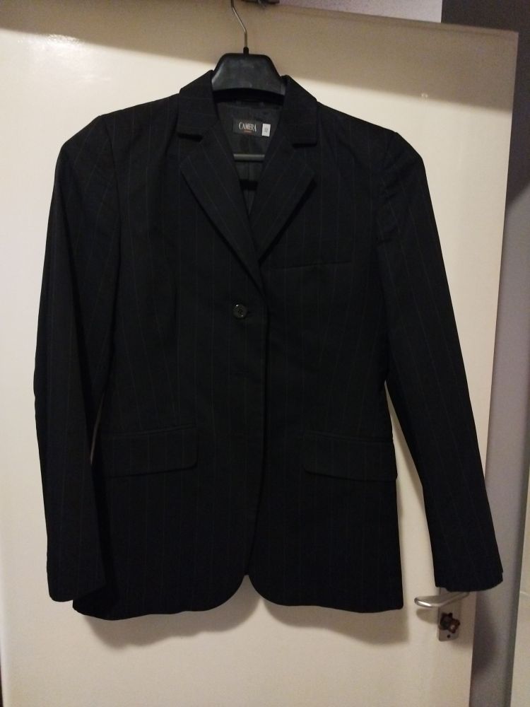 schwarzer Blazer mit Nadelstreifen auf einem Bügel an einer Tür hängend - Vorderseite. 1 Knopf. 1 Brusttasche. 2 Seitentaschen. Kragen bildet V-Ausschnitt. 