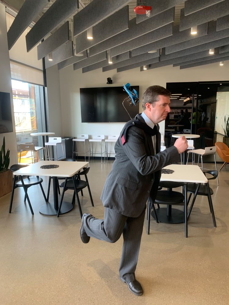 A man in a suit is pretending to run. A small toy plane is suspended by a wire behind him. His tie is swept to the side.