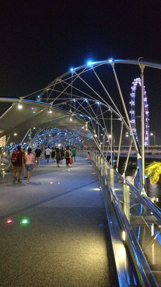 A shot from the Helix Bridge in Singapore. 

Rather than a straight suspension bridge the supports curve over the deck reminiscent of strands of DNA. The parts where the stands meet are lit in different colors. There is a crowd of people walking away along the deck. In the background a giant Ferris wheel is lit up against the night sky.