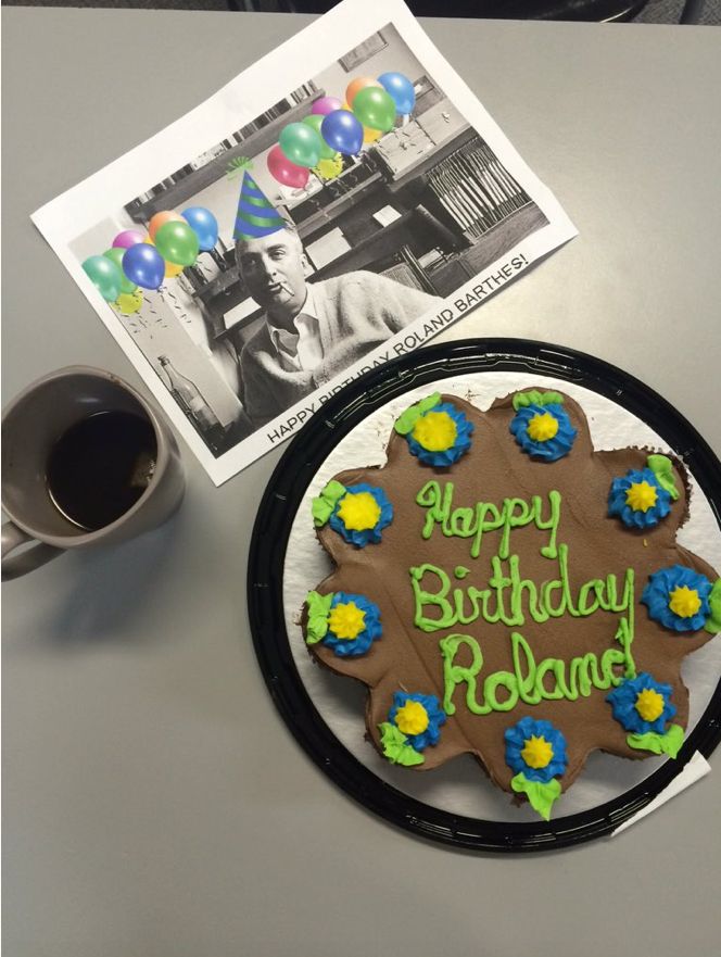 A birthday cake with the inscription, “Happy Birthday Roland” beside a photo of Roland Barthes and a mug full of dark liquid. 