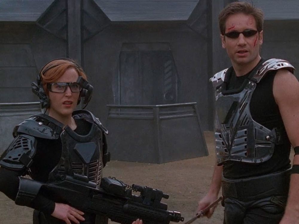 Picture of Mulder and Scully from perhaps the worst episode of the X-Files ever (First Person Shooter) that I post every year on Cyber Monday. Scully is wearing ridiculous body armor, goggles, and holding a big gun. Mulder is wearing a shiny metal chest plate, a sleeveless shirt for some reason, and late 90s-esque tiny sunglasses. He might also be holding a sword? William Gibson what were you thinking
