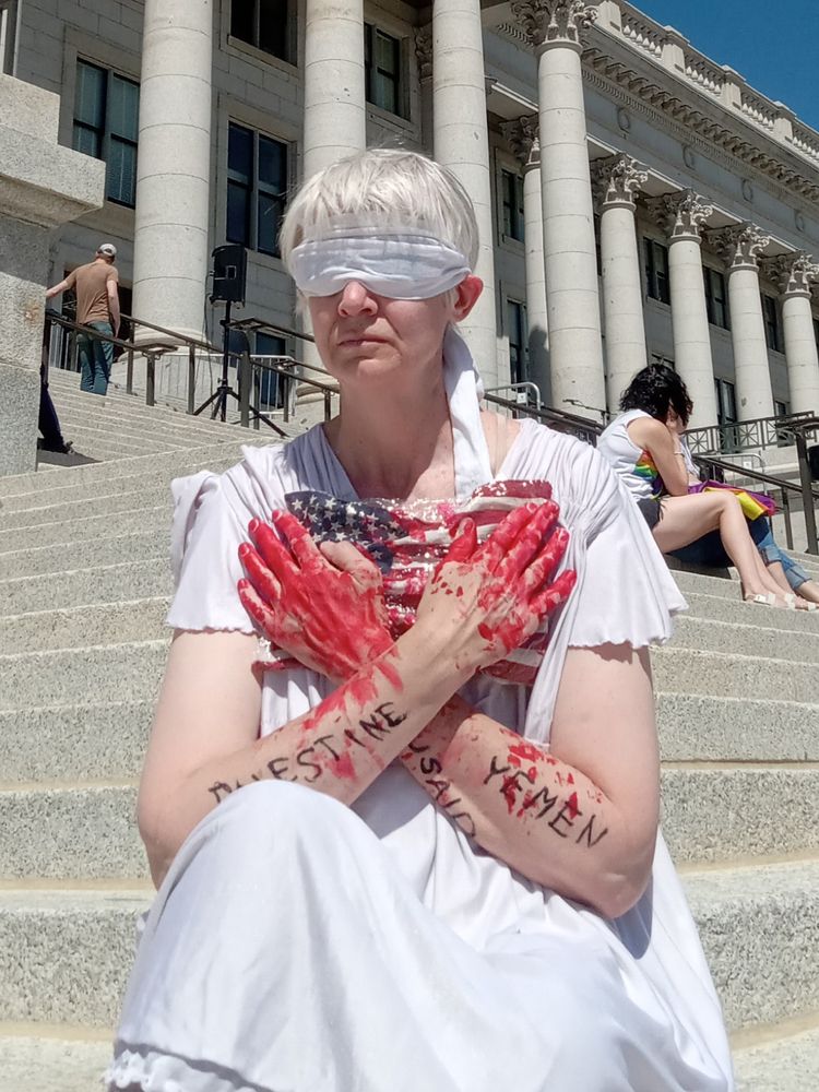 Protestor dressed as justice embraces bloodied U.S. flag with bloodied hands. Palestine, Yemen, and USAID are written on her arms.