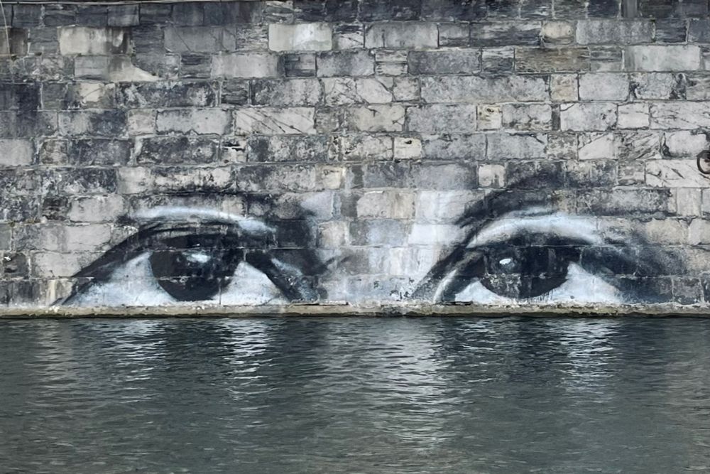 Blick auf ein Stück Mauer an einem Kanal. Direkt auf Höhe der Wasseroberfläche sind zwei Augen gemalt, die einen intensiven Blick haben. Die Augen sind schwarz-weiß gemalt, die Mauer ist aus Steinen in Grautönen, ein Stückchen Wasser, das im Vordergrund zu sehen ist, ist dunkelgrau. 