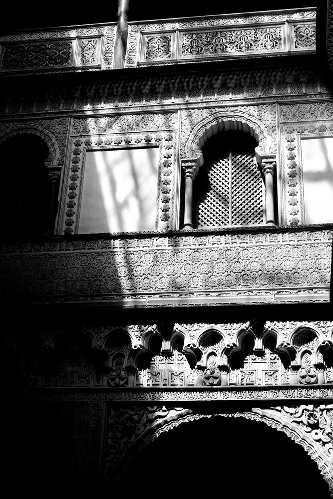Schwarz-weiß Foto. Blick auf ein vergittertes Rundbogenfenster in einem alten Palast. Der Stil ist maurisch beeinflusst. Die Einfassung des Fensters und die Fassade sind mit Steinmetzarbeiten reich verziert. Die Muster sind zum Teil durchbrochen. Es gibt starke Kontraste zwischen Licht und Schatten. 