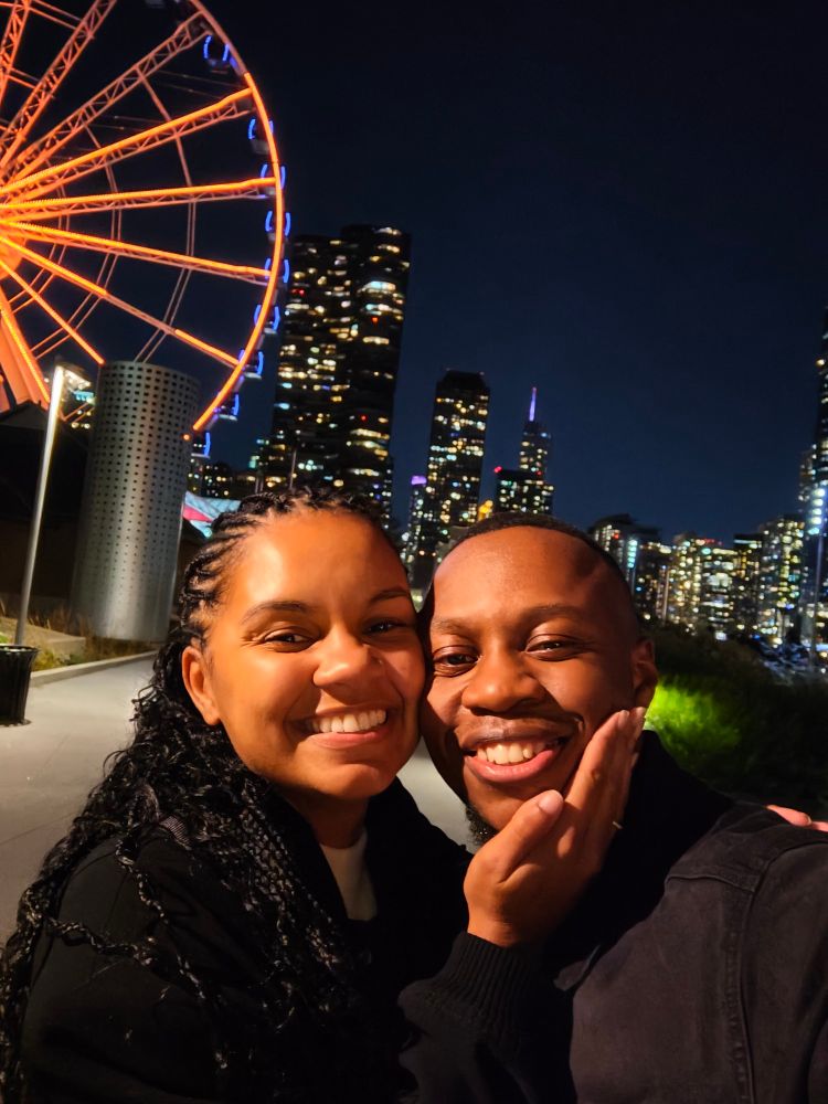 A selfie of Clíona and De-Shaine at Navy Pier in the night. They are in front of an illuminated Ferris wheel and the Chicago Skyline.