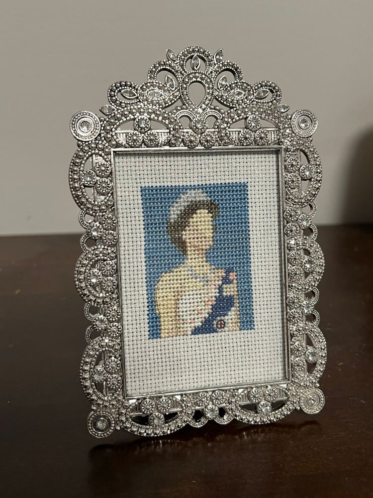 A cross stitch of a woman with a blue sash and silver crown against a blue background in a frame with swirls and white gemstones. 