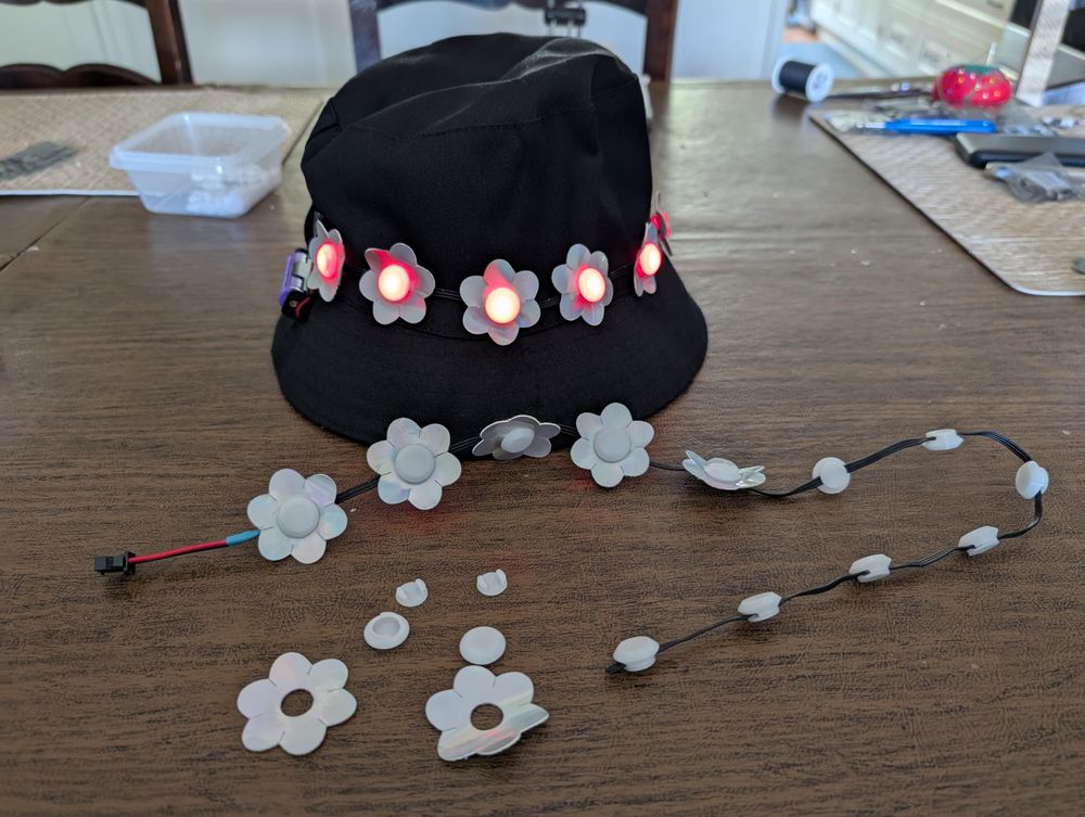 A black bucket cap rests on a table. Several silver flowers with illuminated red centers run around the middle of the hat. In front of the hat is a string of pebble lights in which the LEDs have been covered by translucent circular diffusers. Some of the diffusers form the center for vinyl flowers that have been placed around them. On the table in front of the LED string are two of the 3D printed diffusers, along with two flowers cut from vinyl with holes in their centers for placing the diffusers through.