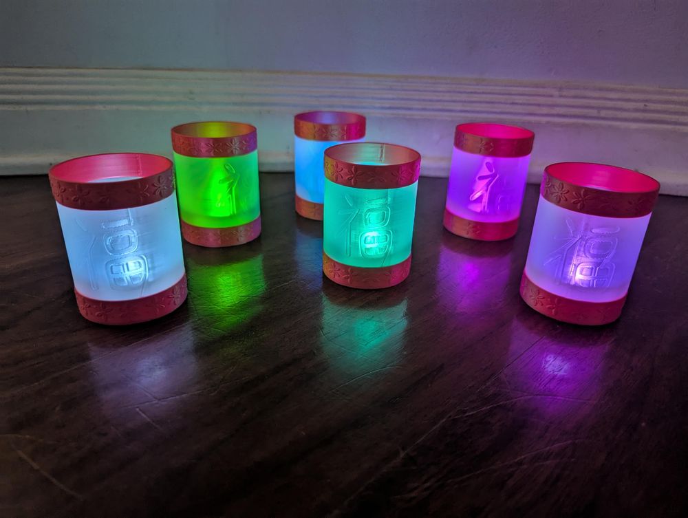 Six of the tea light holders described in the previous image, arranged in an irregular row on a wood floor. Each is illuminated in a different color.