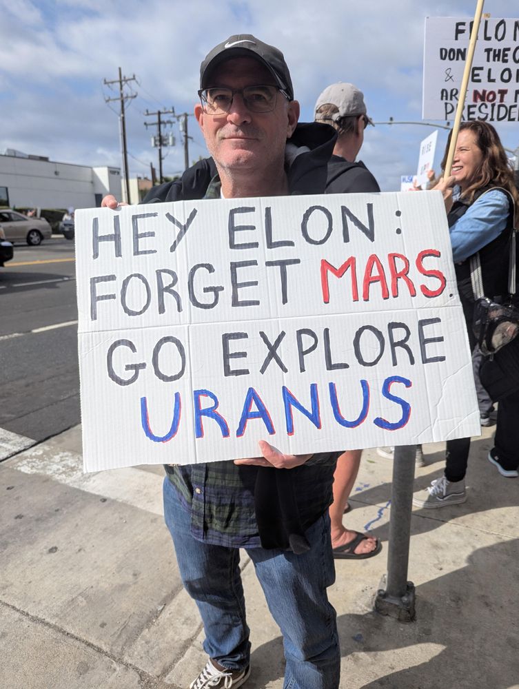 Man carrying a protest sign that reads, "Hey Elon, forget Mars go explore Uranus."