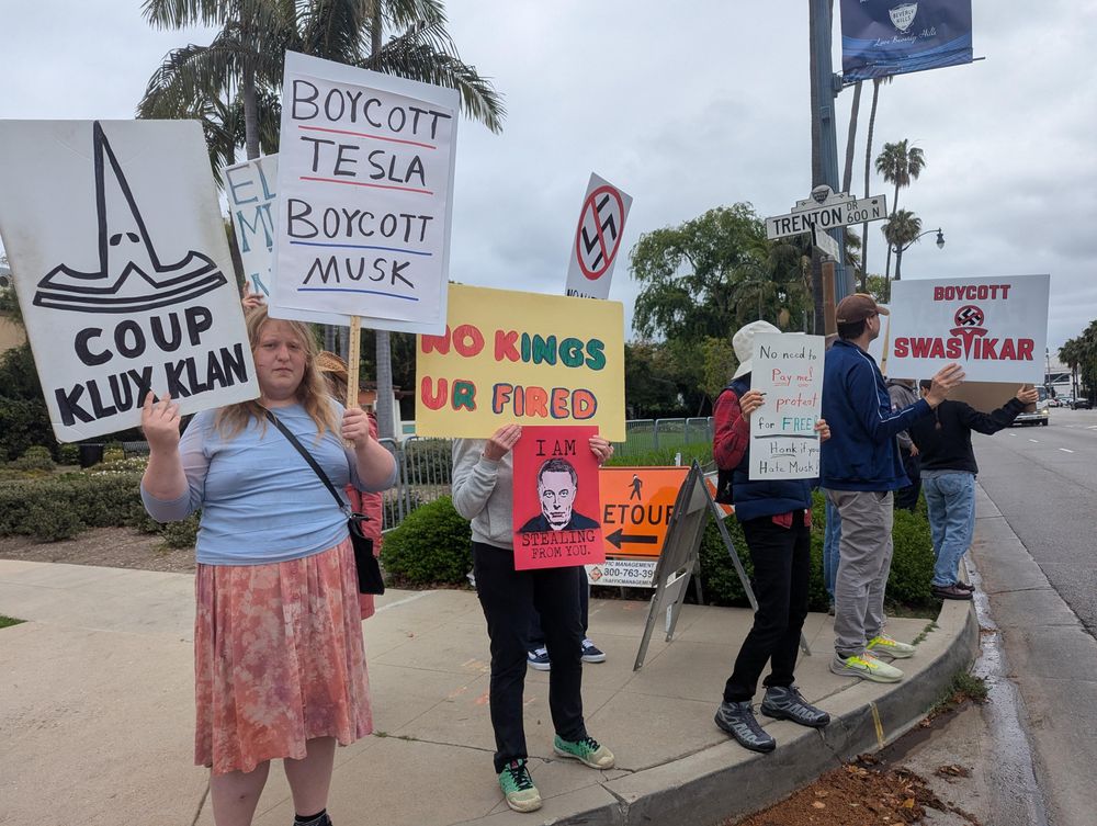 A small group of protesters holding anti-Musk signs across the street from the Beverly Hilton which has a sign announcing the Milken Institute conference 