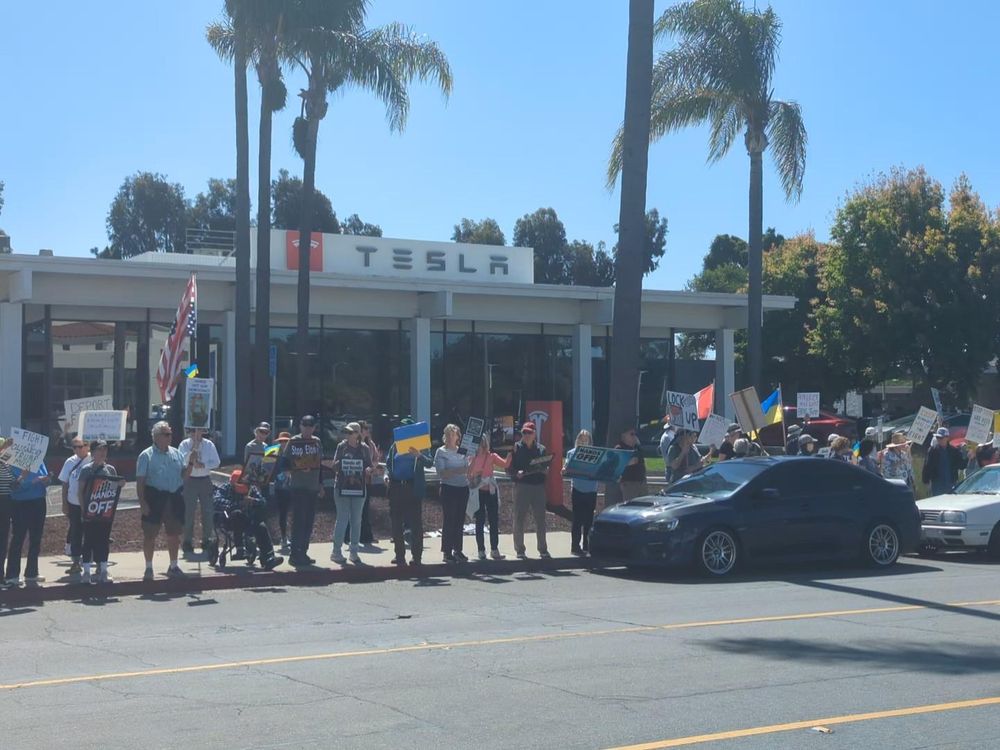 A line of protesters holding signs and standing in front of a Tesla dealership 