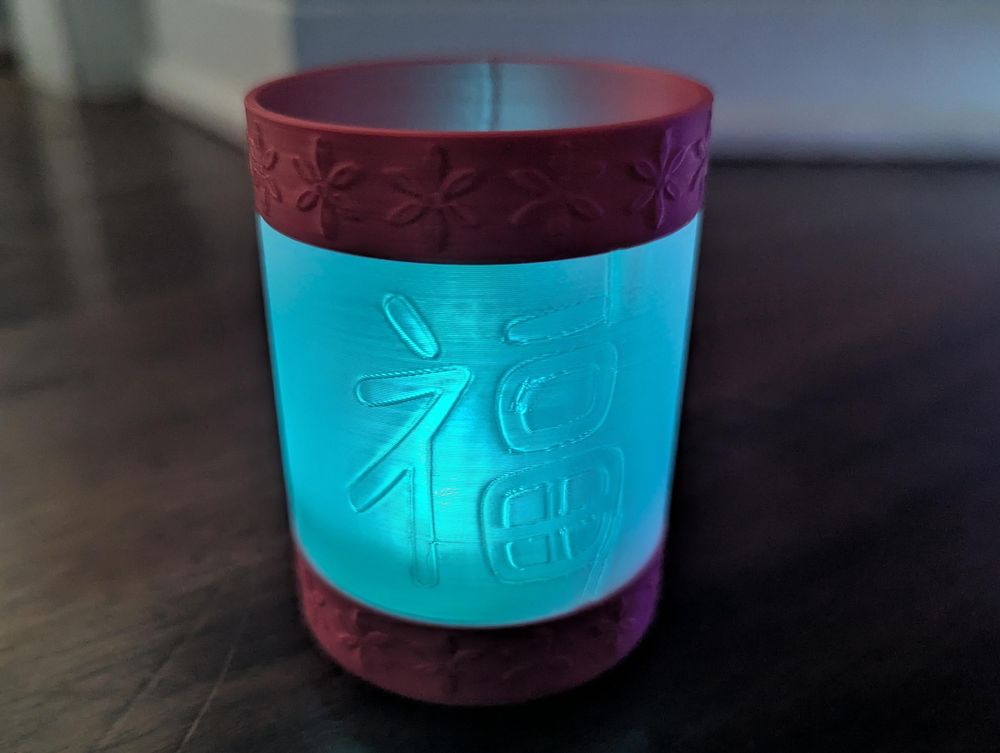 Close up of an illuminated cylindrical 3d printed tea light holder with a red top and bottom rim about 1/2 inches in height embossed with a floral design and a translucent middle section about 2 inches in height embossed with the Chinese character for good luck. The light inside the holder is glowing blue.
