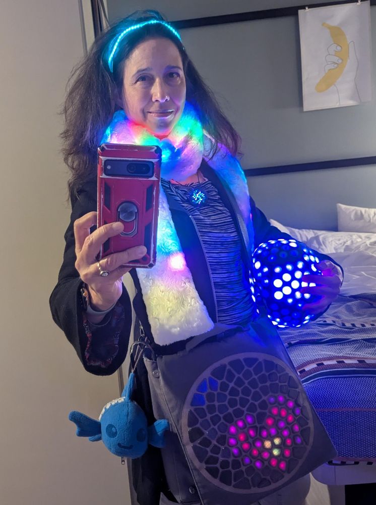 Me, taking a selfie in a mirror wearing an LED headband, illuminated Lux Lavalier necklace, a white LED scarf, carrying a blue soft icosahedron plush studded with LED circles. Also carrying a messenger back with an illuminated voronoi pattern displaying a heart,.and attached to the messenger bag is a blie plush dish with illuminated scales.