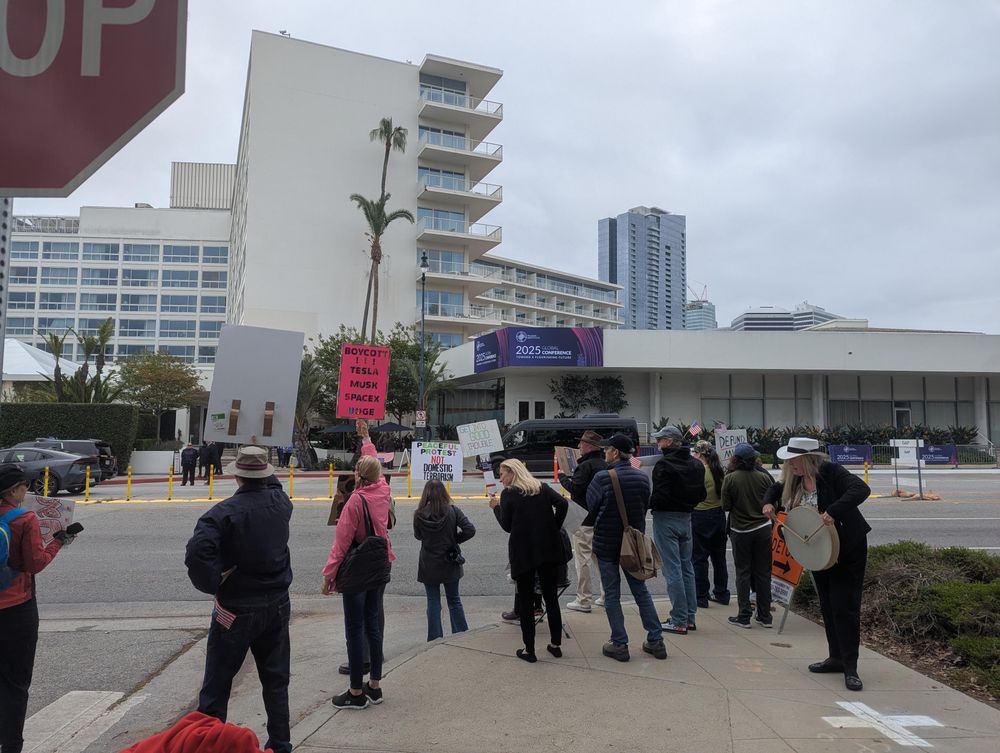 A small group of anti Musk protestors on a street corner holding signs