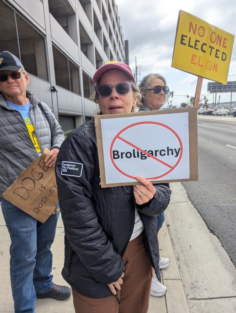 A woman carrying a white sign with a circle and red slash through the word, "broligarchy"